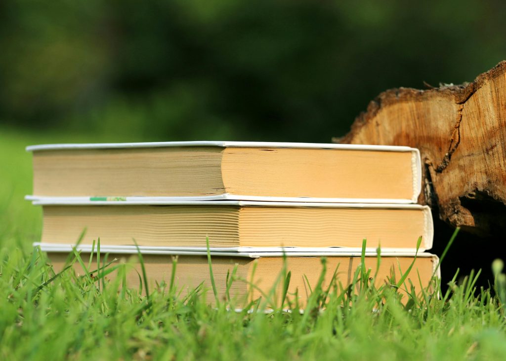 Three books stacked on grass next to a tree log in a peaceful outdoor setting.